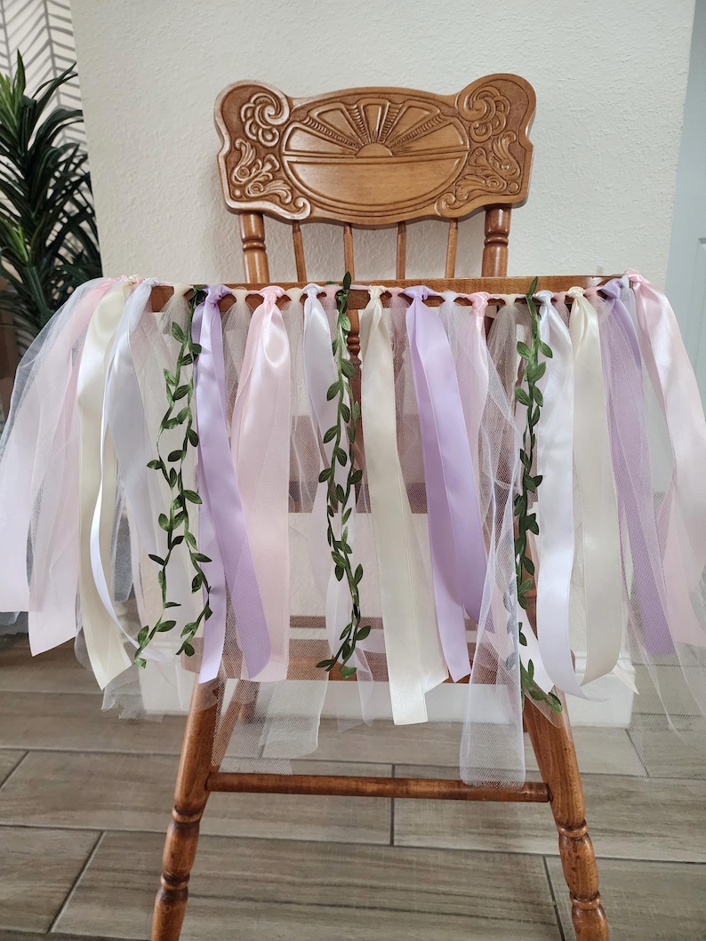 May include: A wooden chair decorated with a tulle skirt and ribbons in shades of pink, white, and lavender. Green vine accents are interspersed among the ribbons, creating a whimsical and festive look. The chair has a decorative back.