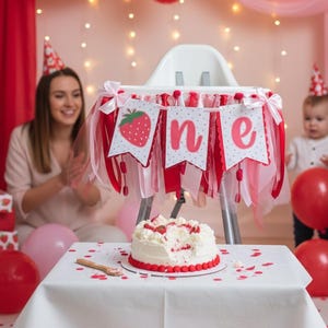 May include: A high chair decorated with a white banner featuring a red strawberry and the text "one" in pink letters, accented with red and pink fabric ribbons and bows. A white frosted cake with red strawberry pieces sits on a white table.