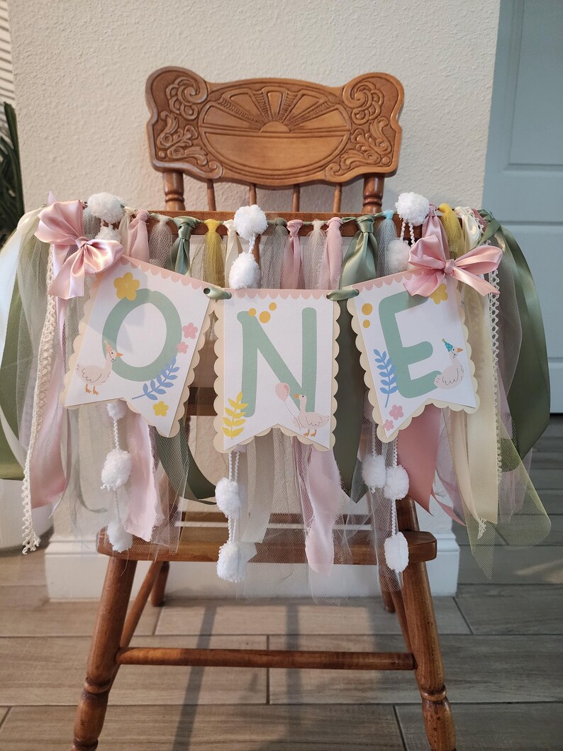 May include: A 'ONE' banner for a first birthday celebration. The banner is attached to a wooden high chair. The banner has three scalloped-edged panels with the letters 'O', 'N', and 'E'. Each panel features a duck illustration and floral accents. The banner is decorated with pink bows, tulle, and white pom-poms.