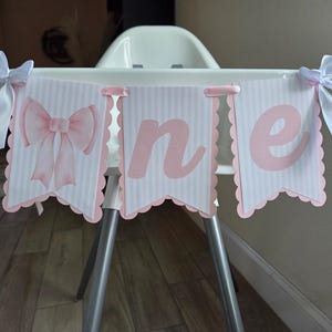 May include: A pink and white banner with the word "one" displayed on a high chair. The banner has a scalloped edge and is decorated with a pink bow. White ribbons are tied to the high chair.