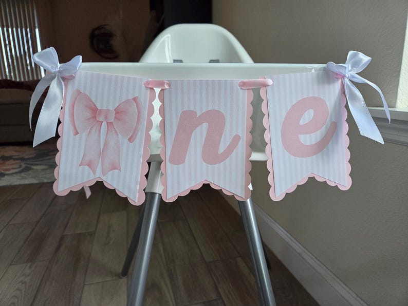 May include: A pink and white banner with the word "one" displayed on a high chair. The banner has a scalloped edge and is decorated with a pink bow. White ribbons are tied to the high chair.
