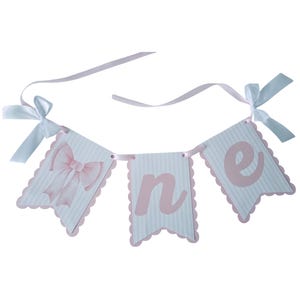 May include: A banner with three pennants spelling "one" in pink letters. The pennants are white with pink stripes and scalloped edges. A pink bow is on the first pennant. White ribbons with bows are attached.
