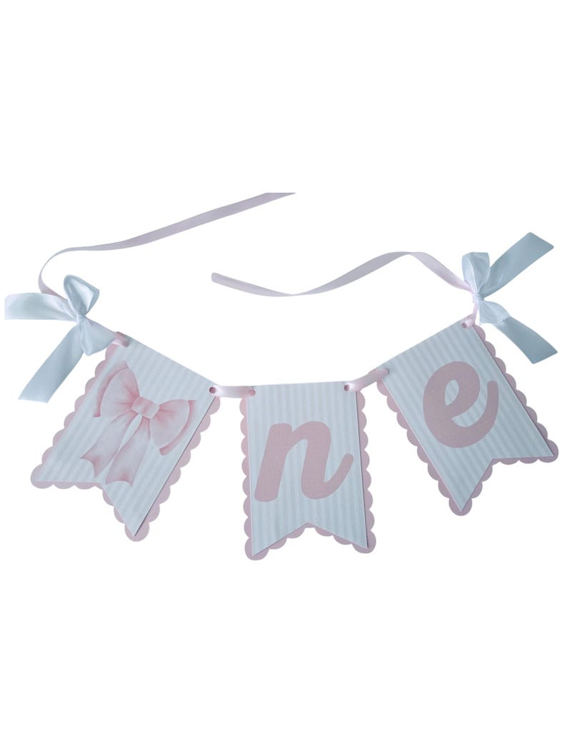 May include: A banner with three pennants spelling "one" in pink letters. The pennants are white with pink stripes and scalloped edges. A pink bow is on the first pennant. White ribbons with bows are attached.