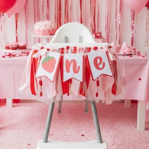 May include: A high chair banner featuring three white pennants with red scalloped edges and a strawberry graphic, followed by the letters n and e in red script. The banner is decorated with layers of red, white, and pink tulle and ribbon.
