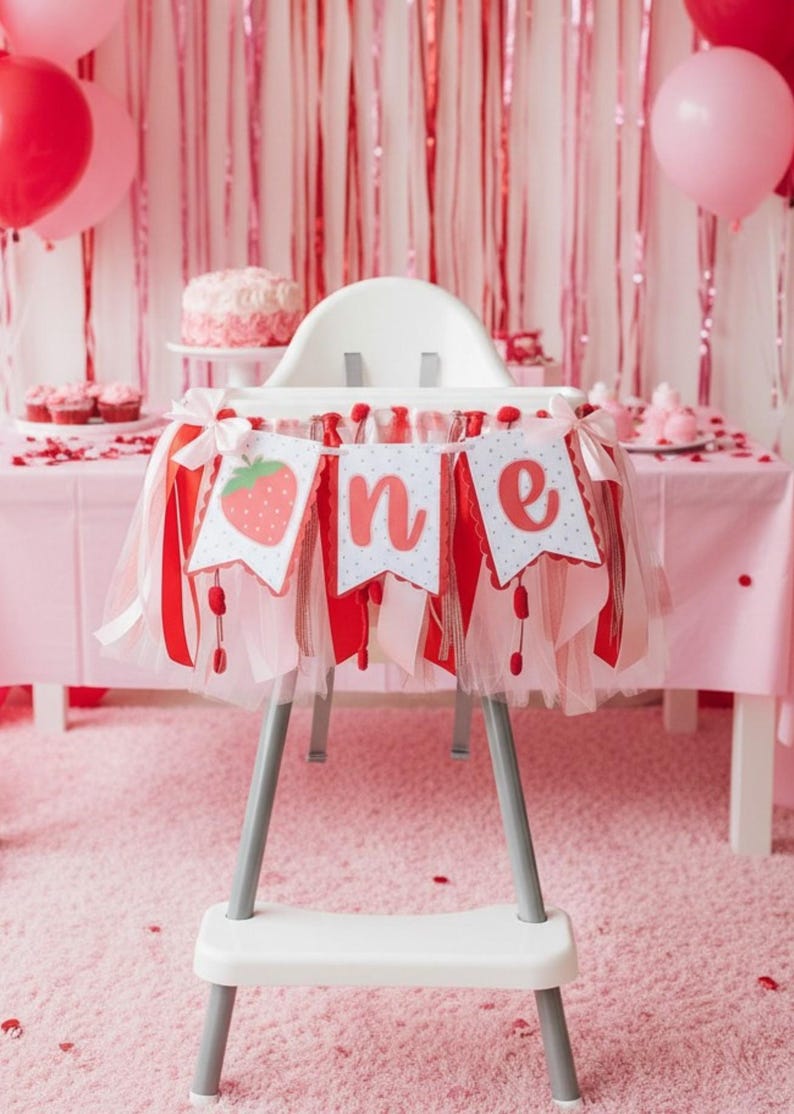May include: A high chair banner featuring three white pennants with red scalloped edges and a strawberry graphic, followed by the letters n and e in red script. The banner is decorated with layers of red, white, and pink tulle and ribbon.