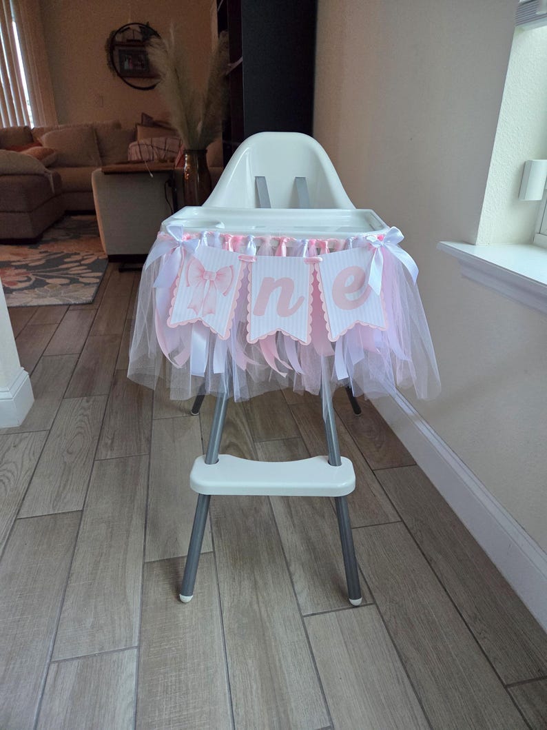 May include: A white high chair decorated with a pink and white tulle skirt and banner that reads "one". The banner has pink bows and scalloped edges. The high chair has a metal frame and is set on a light wood floor.