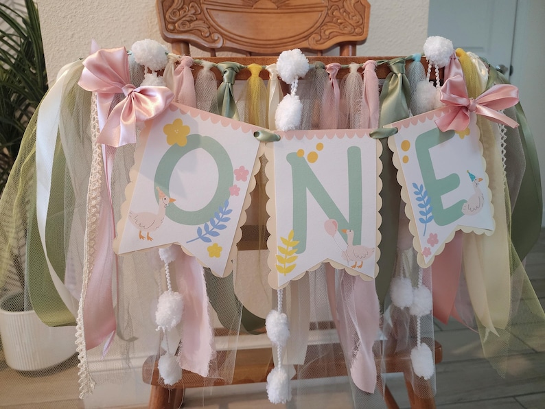 以下が含まれることがあります： パステルグリーンの文字で「ONE」と書かれた装飾バナー。ガチョウのイラストと花のアクセントで飾られています。ピンクのリボン、白いポンポン、リボンで装飾され、お祝いの雰囲気を醸し出しています。