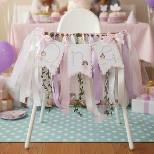 May include: A high chair decorated for a first birthday party. The chair is adorned with a banner that says "one" and has ribbons in white, pink, and lavender. Balloons, gifts, and a table with a pink tablecloth are in the background.