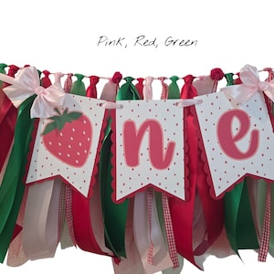 May include: A decorative banner featuring three white pennants with scalloped edges and red polka dots. The pennants display a strawberry illustration and the letters O, N, and E in pink. The banner is adorned with hanging pink, red, and green ribbons.