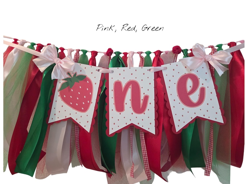 May include: A decorative banner featuring three white pennants with scalloped edges and red polka dots. The pennants display a strawberry illustration and the letters O, N, and E in pink. The banner is adorned with hanging pink, red, and green ribbons.