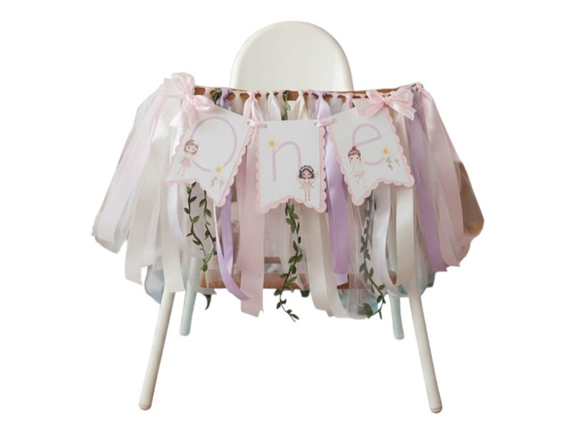 May include: A high chair decorated with a pink, white, and lavender tulle skirt and ribbon streamers. The banner reads "ONE" with illustrations of a fairy. Greenery accents the banner and skirt, creating a whimsical look.