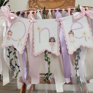 May include: A decorative banner with the word "one" spelled out on three scalloped-edge pennants. Each pennant features a fairy illustration and floral accents. The banner is adorned with pink, white, and lavender ribbons and tulle.