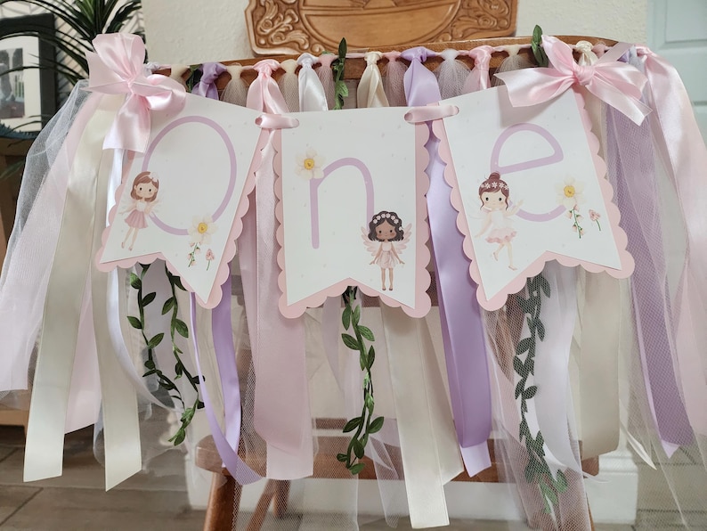 May include: A decorative banner with the word "one" spelled out on three scalloped-edge pennants. Each pennant features a fairy illustration and floral accents. The banner is adorned with pink, white, and lavender ribbons and tulle.