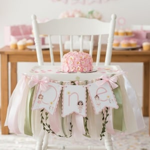 May include: A white high chair decorated for a first birthday. The chair has a pink and white tutu skirt with bows and a banner that says "one". A small pink cake sits on the tray. Cupcakes and a table are in the background.