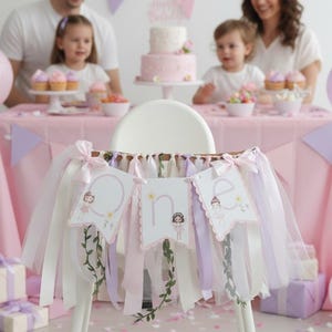 May include: A high chair decorated with a banner that says "One" with a ballerina design. The banner is decorated with ribbons in shades of pink, white, and lavender. A pink tablecloth, balloons, and a cake are in the background.