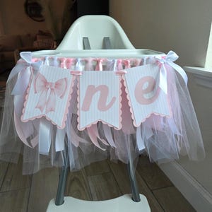 May include: A high chair decorated for a first birthday. The chair is adorned with a banner that spells "one" in pink letters on white pennants. Pink and white tulle and ribbons are draped around the chair, creating a festive look.