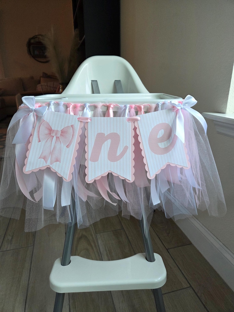 May include: A high chair decorated for a first birthday. The chair is adorned with a banner that spells "one" in pink letters on white pennants. Pink and white tulle and ribbons are draped around the chair, creating a festive look.