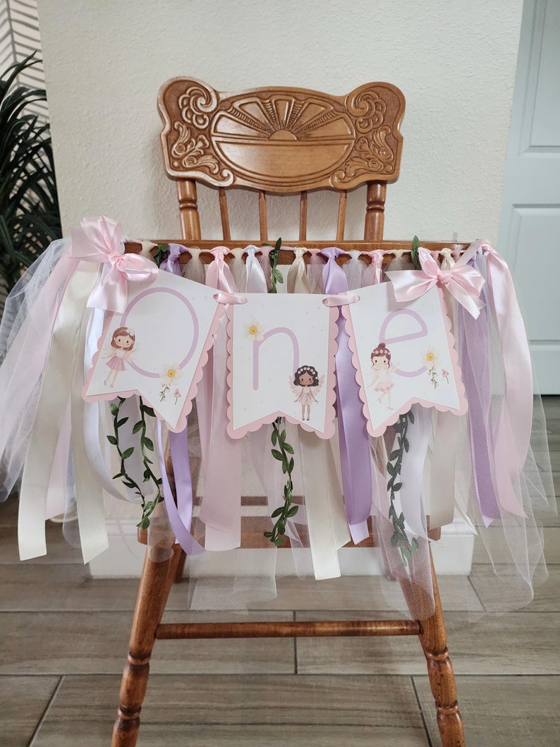 May include: A high chair banner with the word "one" spelled out on three pink and white pennants. The banner is decorated with ribbons, tulle, and floral accents. The chair is made of wood and has a decorative back.