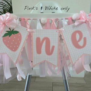 May include: A high chair banner featuring three pennants with a light pink and white gingham pattern. The pennants display a strawberry graphic and the letters O, N, and E in pink. The banner is decorated with pink satin bows and tulle.