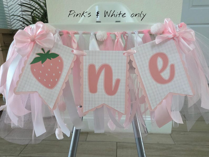 May include: A high chair banner featuring three pennants with a light pink and white gingham pattern. The pennants display a strawberry graphic and the letters O, N, and E in pink. The banner is decorated with pink satin bows and tulle.