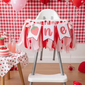 May include: A white high chair decorated with a strawberry-themed banner featuring the word one in red script. The banner is adorned with red and pink tulle ribbons and bows. The high chair is surrounded by red and pink balloons and strawberry decor.