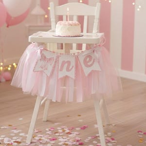 May include: A white high chair decorated for a first birthday. A small cake with a lit candle sits on the tray. A pink tulle skirt and a banner that says "one" adorn the chair. Pink balloons and confetti complete the festive scene.