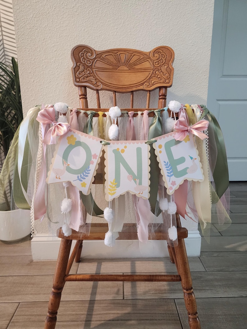 May include: A wooden high chair decorated for a first birthday celebration. A banner that spells 'ONE' is draped across the front, with each letter on a scalloped-edge card. The banner is adorned with ribbons, bows, and pom-poms in shades of pink, green, and white.