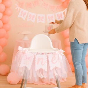 May include: A high chair decorated for a first birthday celebration. The chair has a pink tulle skirt and a banner that says "one". Pink balloons and a cake are in the background.