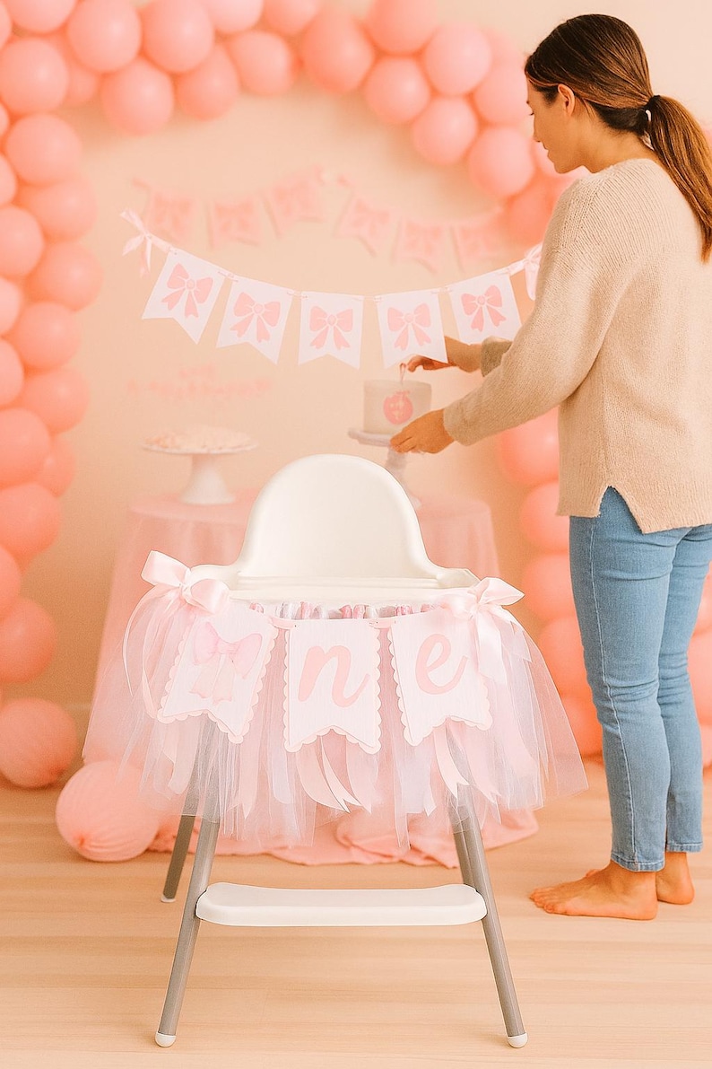 May include: A high chair decorated for a first birthday celebration. The chair has a pink tulle skirt and a banner that says "one". Pink balloons and a cake are in the background.