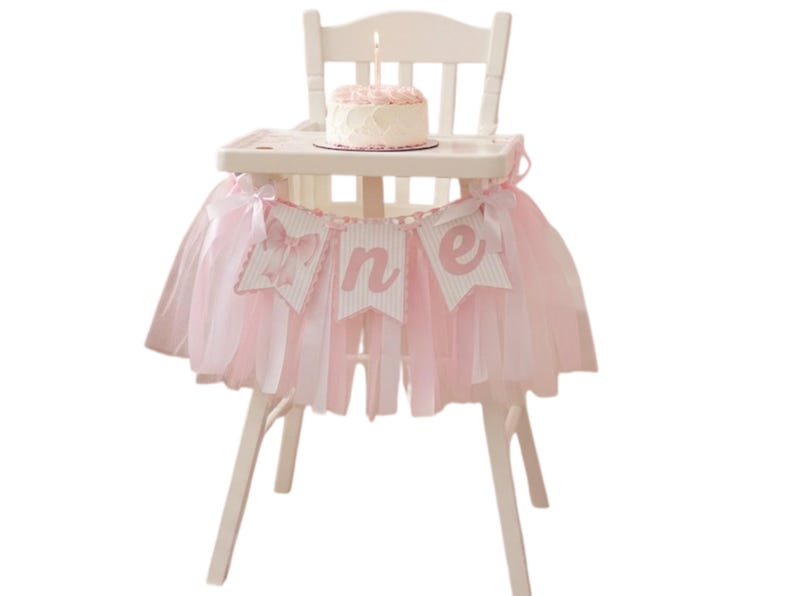 May include: A white high chair decorated with a pink tutu skirt and a banner that says "one". A small cake with a lit candle sits on the tray. The chair is set against a white background.