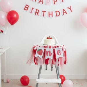 May include: A white high chair decorated with a red and white tulle skirt and a banner featuring a strawberry and the word one. A small white cake with a number one topper sits on the tray. A Happy Birthday banner hangs on the wall above.