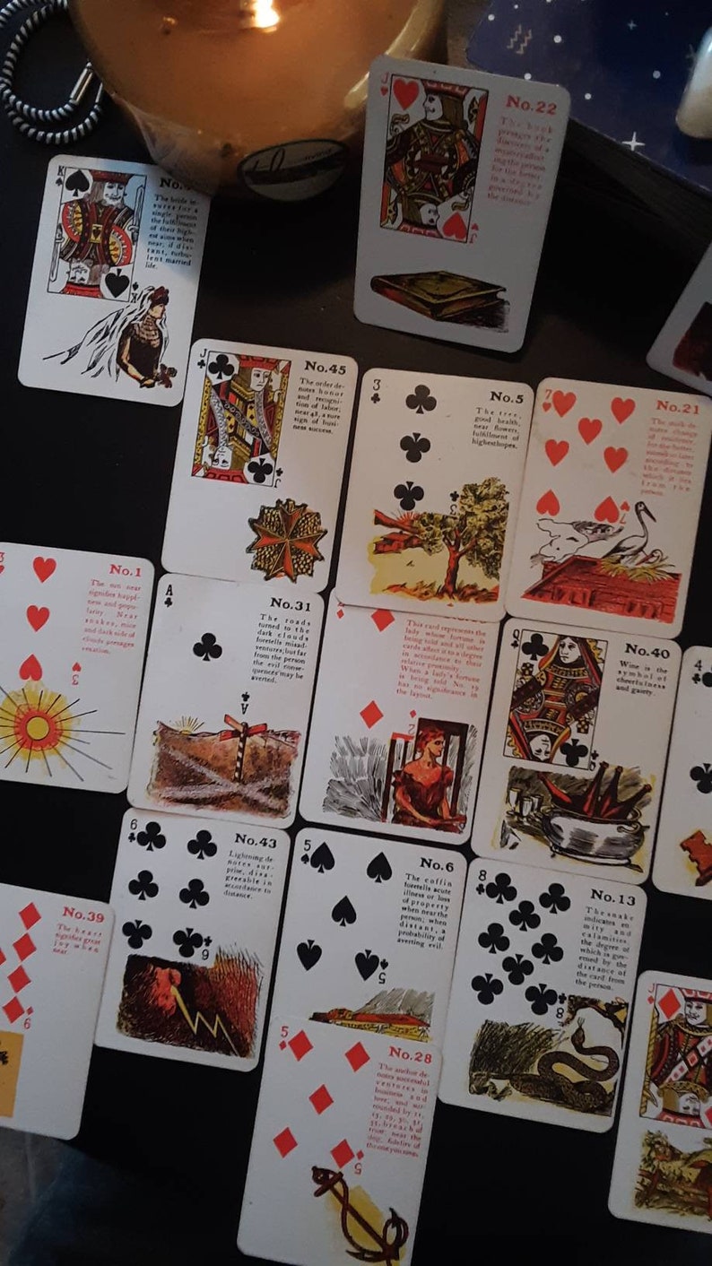 Op de afbeelding: Een set vintage speelkaarten met kleurrijke illustraties en tekstbeschrijvingen. De kaarten zijn genummerd en bevatten verschillende symbolen, waaronder een zon, een boom, een weg, een slang en een vogel. De kaarten liggen op een donker oppervlak.