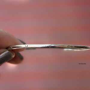 May include: A silver bangle bracelet with a simple design featuring a circle, an X, and a circle pattern.