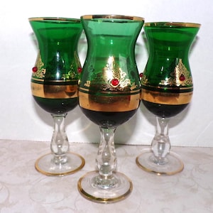 May include: Three green glass goblets with gold trim and ornate gold leaf designs. Each goblet has a clear stem and base with gold trim, and a small red jewel. The goblets are decorative.