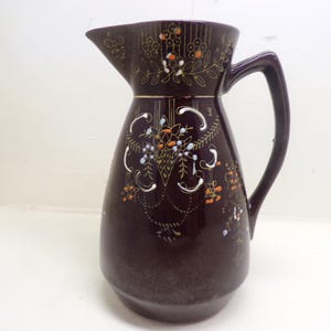 May include: A dark brown ceramic pitcher with a decorative floral design in white, orange, and blue. The pitcher has a curved handle and a spout for pouring. The floral design is on the front and top of the pitcher.