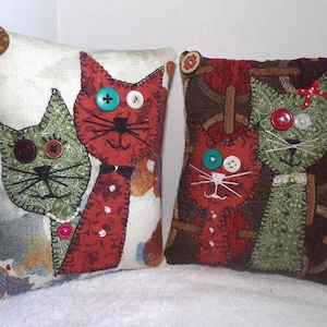May include: Two decorative cat-themed pillows. One pillow has a cream background with a floral pattern and a green and red cat applique. The other pillow has a brown background with a red and green cat applique. Both cats have button eyes.