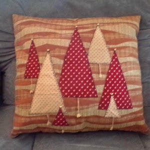 Puede incluir: Un cojín decorativo con un fondo a rayas marrones y beige. Presenta árboles de Navidad aplicados con diseños de estrellas rojas y lunares crema. Los árboles están cosidos con hilo blanco y tienen pequeños detalles de cuentas blancas.