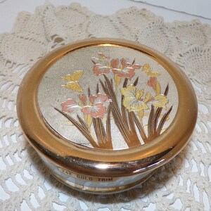May include: A round, silver trinket box with a gold trim and a floral design featuring pink and yellow flowers and butterflies. The box has the text "24K GOLD TRIM" on the bottom.