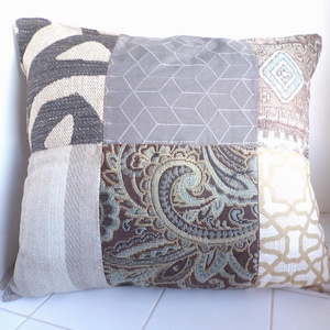 May include: Square patchwork pillow with various fabric patterns. The pillow features sections of gray geometric, zebra print, and paisley designs. The color palette includes gray, beige, brown, and teal.