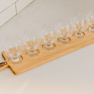 Op de afbeelding: Een houten bierproefplank met zes lege glazen. De plank heeft een handvat met de tekst "Tin Roof" erin gebrand.