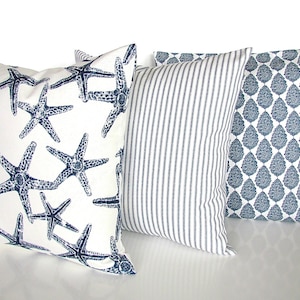 May include: Three decorative throw pillows with a nautical theme. The first pillow has a white background with a navy blue starfish pattern. The second pillow has a white background with navy blue stripes. The third pillow has a white background with a navy blue paisley pattern.