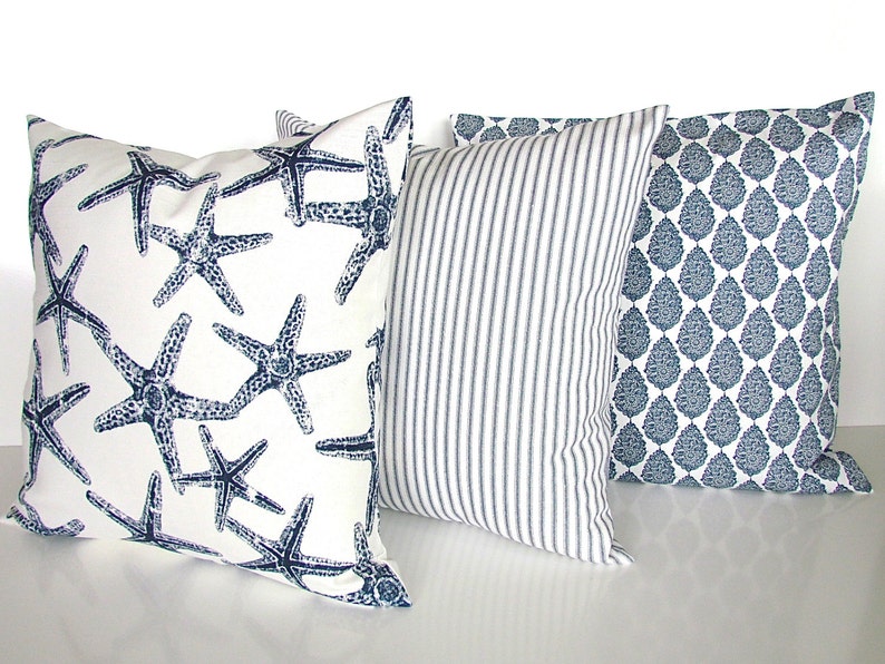 May include: Three decorative throw pillows with a nautical theme. The first pillow has a white background with a navy blue starfish pattern. The second pillow has a white background with navy blue stripes. The third pillow has a white background with a navy blue paisley pattern.