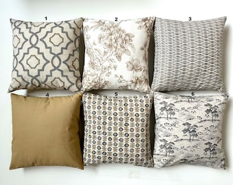 May include: Six square decorative throw pillows. The pillows feature various patterns, including geometric, floral, and landscape designs. Colors include neutral tones of beige, brown, and gray. One pillow is a solid tan color.