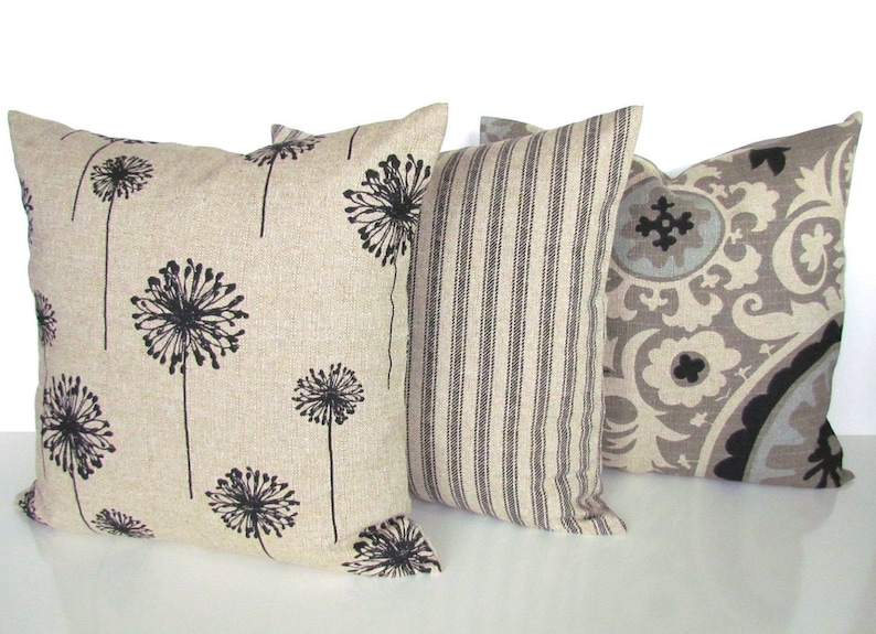 May include: Three decorative throw pillows with a neutral color scheme. One pillow features a black and white dandelion print, another has black and white stripes, and the third has a black, gray, and white paisley pattern.