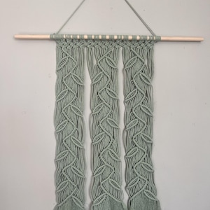 May include: A sage green macrame wall hanging featuring three vertical panels with a repeating leaf-like knot pattern. The cotton cord is suspended from a light-colored wooden dowel with a matching cord loop for hanging on the wall.