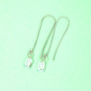 May include: A pair of silver-toned drop earrings. Each earring features a long, curved wire that hangs from a small, round bead. A small ring connects the wire to a rectangular, iridescent crystal. The earrings are set against a light green background.
