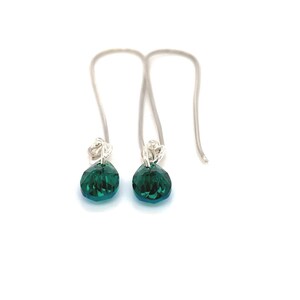 May include: A pair of dangle earrings featuring faceted, teardrop-shaped, emerald green crystals. The crystals are suspended from silver-colored, curved ear wires. The earrings are displayed against a white background.