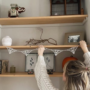 May include: A person hangs a white spiderweb garland on a wooden shelf with various decorative items, including a ceramic elephant, a birdcage, and framed pictures.
