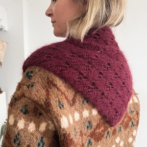 May include: A burgundy knit cowl neck scarf with a geometric pattern is worn over a brown and tan patterned sweater. The sweater has white and teal accents. The cowl neck is a deep burgundy color.