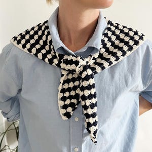 Könnte beinhalten: Schwarz-cremefarbener gehäkelter Schal mit Karomuster. Der Schal ist geknotet und über einem hellblauen Button-Down-Hemd getragen. Der Schal ist ein stilvolles Accessoire.